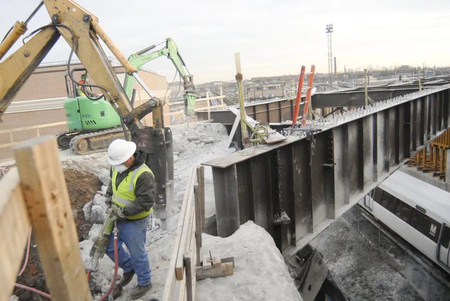 Laborer breaking concrete with a jackhammer beside steel girders over a rail line